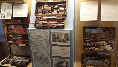 Display of vintage surgical instruments in wooden cases, with historical information and illustrations on the wall.