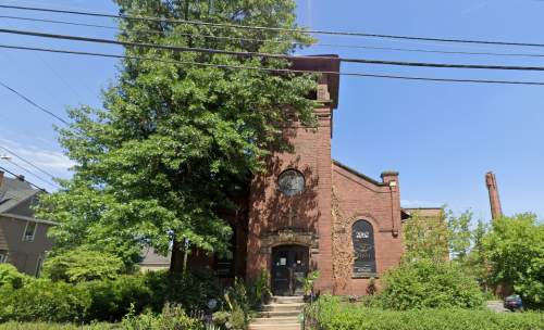 A red brick building with large windows, surrounded by greenery and trees, under a clear blue sky.