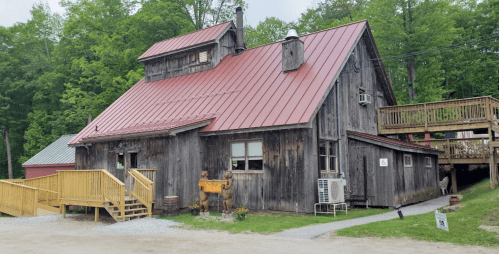 A rustic wooden building with a red metal roof, featuring a porch and surrounded by greenery.