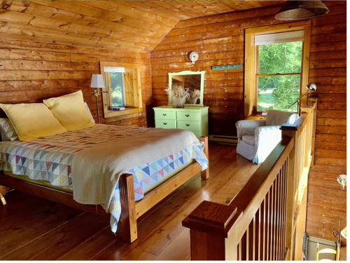 Cozy wooden bedroom with a quilted bed, green dresser, and a chair, featuring large windows and warm lighting.