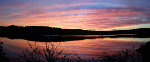 A serene lake at sunset, reflecting vibrant pink and purple clouds, with silhouettes of trees and sailboats in the distance.