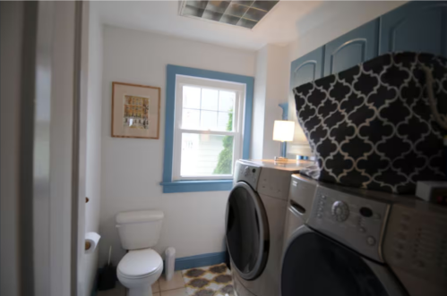A bright laundry room featuring a washer and dryer, a toilet, and blue accents on the walls and cabinets.