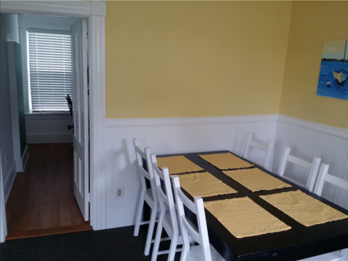 A bright dining area with a black table set for eight, yellow placemats, and a doorway leading to another room.