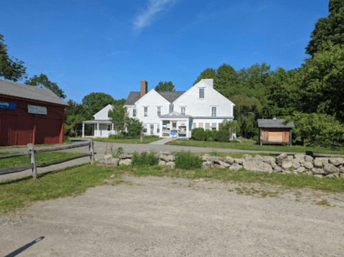 A white house with a porch surrounded by greenery, near a gravel path and a red barn in the background.