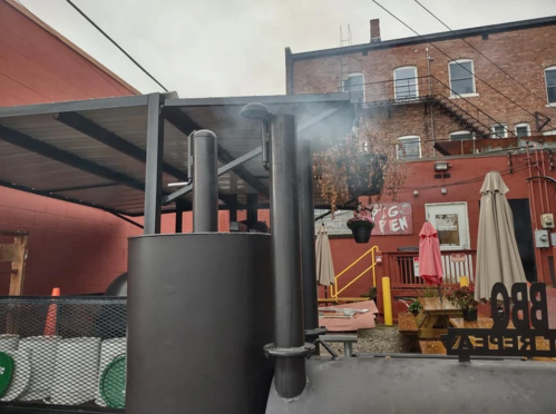 A BBQ smoker with smoke rising, set against a red brick building and outdoor seating area.