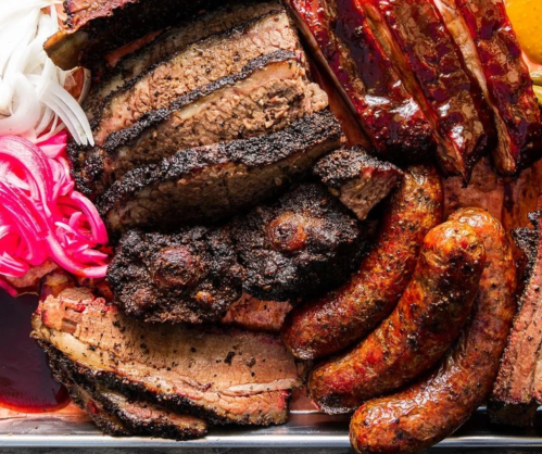 A platter of smoked meats including brisket, sausage, and ribs, garnished with pickled onions and a slice of orange.