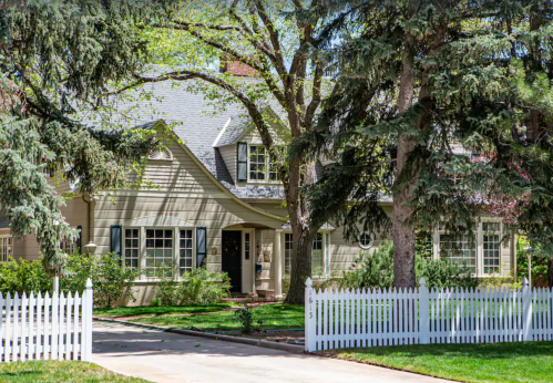 A charming house with a gray roof, surrounded by trees and a white picket fence, set in a lush green landscape.