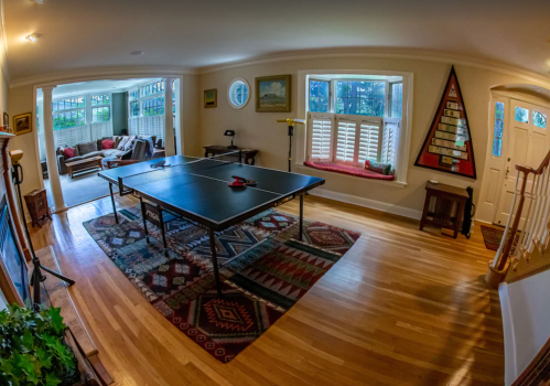 A spacious room with a ping pong table, hardwood floors, and cozy seating area visible through an open doorway.