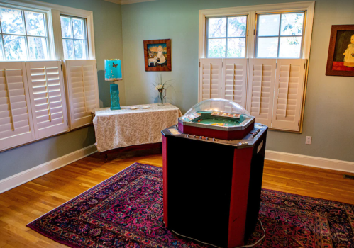 A vintage jukebox sits in a cozy room with large windows, a table, and decorative art on the walls.