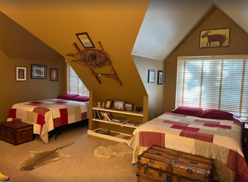 Cozy attic bedroom with two beds, wooden decor, and a bookshelf, featuring warm tones and natural light.