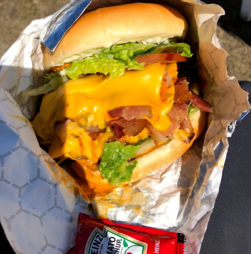 A close-up of a cheeseburger with lettuce, bacon, and melted cheese, wrapped in paper, alongside a ketchup packet.