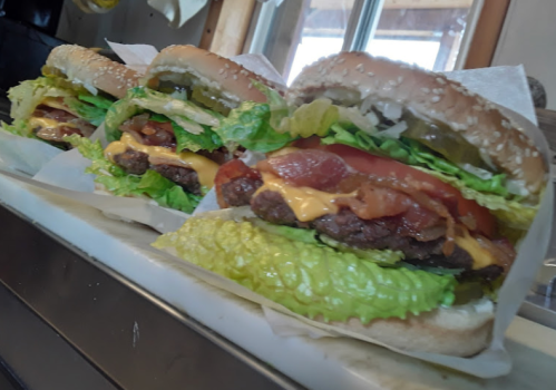 Three large burgers with sesame seed buns, lettuce, tomato, cheese, bacon, and pickles, arranged on a tray.