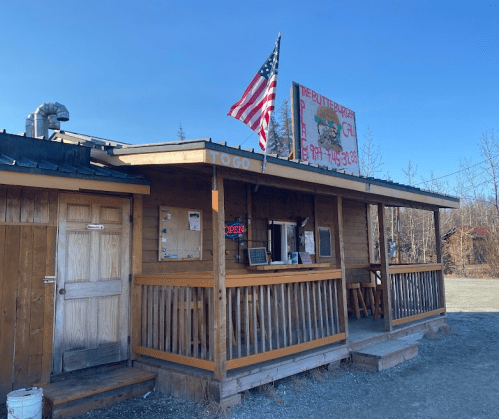 A rustic wooden building with a porch, an American flag, and a sign for a burger joint, open for takeout.