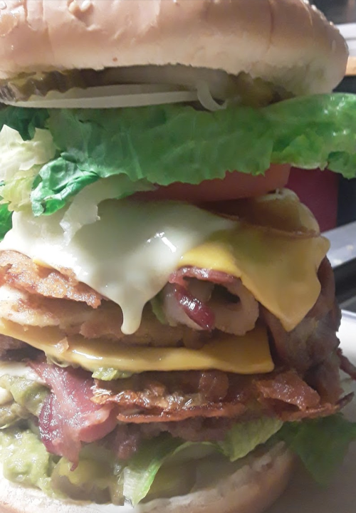 A towering burger with layers of lettuce, tomato, cheese, bacon, and grilled chicken, all in a soft bun.