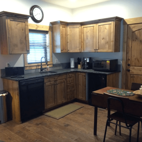 A cozy kitchen with wooden cabinets, a black sink, and appliances, featuring a small table and clock on the wall.
