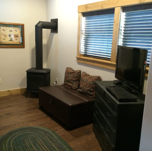 Cozy room with a brown couch, a black TV, a wood stove, and windows with blinds. Rustic decor and wooden accents.