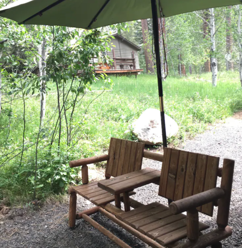 A wooden bench with an umbrella sits on a gravel path, surrounded by greenery and a cabin in the background.