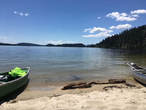 Two canoes on a sandy beach by a calm lake, surrounded by trees and distant hills under a clear blue sky.