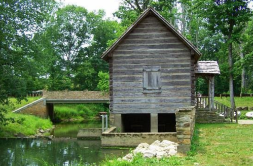 A rustic wooden building beside a calm stream, with a small bridge and lush greenery in the background.
