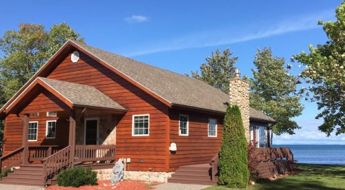 A cozy wooden house with a stone chimney, surrounded by trees and overlooking a calm lake under a clear blue sky.