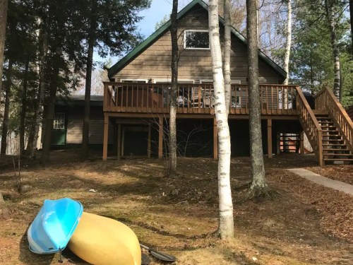 A wooden cabin surrounded by trees, with a deck and two colorful kayaks resting on the ground.