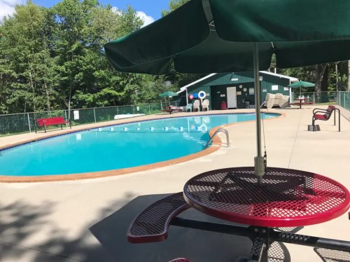 A clear swimming pool surrounded by lounge chairs and shaded tables, set in a green, wooded area.