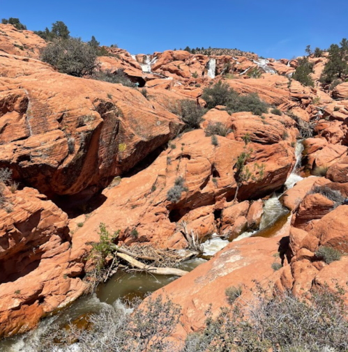 A scenic view of red rock formations with a flowing stream and sparse vegetation under a clear blue sky.