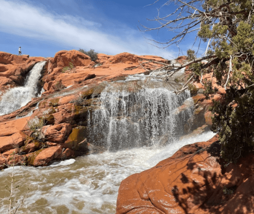 A waterfall cascades over red rock formations under a blue sky, surrounded by sparse vegetation.