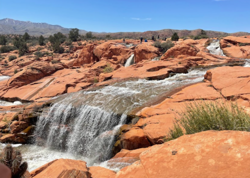 A cascading waterfall flows over vibrant orange rock formations under a clear blue sky.