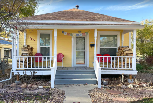 A yellow house with a porch, two red chairs, and stacked firewood on either side, surrounded by a garden.