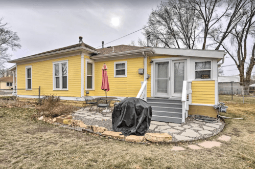 A yellow house with a patio, table, and grill, surrounded by grass and trees under a cloudy sky.