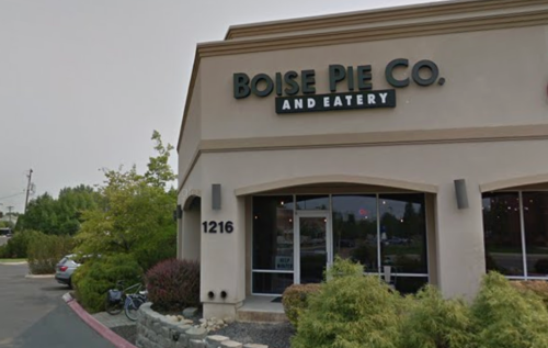 Exterior of Boise Pie Co. and Eatery, featuring a sign and large windows, with greenery and parked cars nearby.