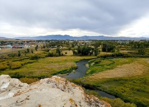 A scenic view of a river winding through fields, with mountains and a small town in the background under a cloudy sky.