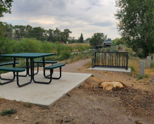 A picnic table on a concrete pad beside a path, surrounded by greenery and trees under a cloudy sky.