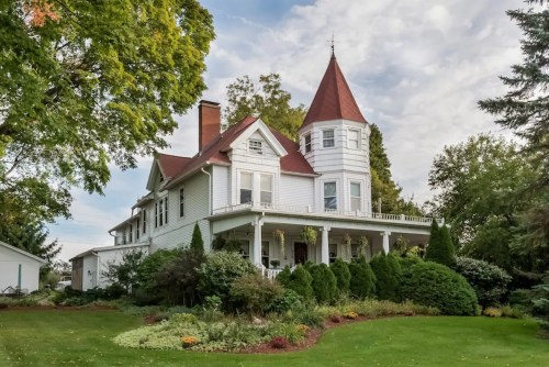 A charming white Victorian house with a red turret, surrounded by lush greenery and colorful landscaping.