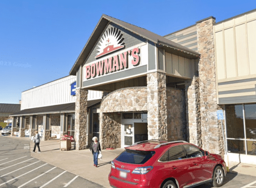 Exterior of Bowman's store with stone facade, clear blue sky, and a red car parked in front. Shoppers are visible.