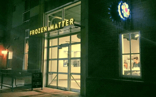 Exterior of "Frozen Matter" shop at night, featuring large glass doors and a colorful sign.