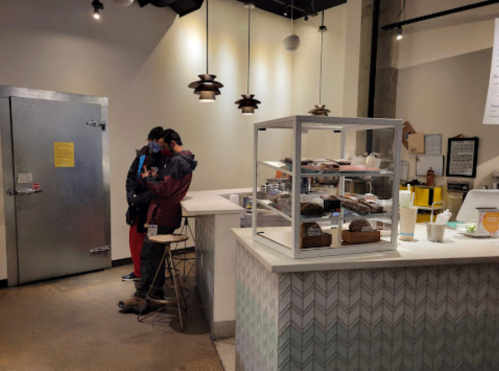 Two people in masks stand at a counter in a cafe, with a display case of baked goods and a large metal door in the background.