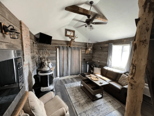 Cozy living room with rustic decor, a leather sofa, antler accents, and a wood stove, featuring large windows.