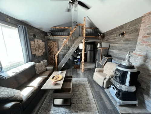 Cozy living room with a sofa, armchair, wooden stairs, and a vintage stove, featuring rustic decor and natural light.