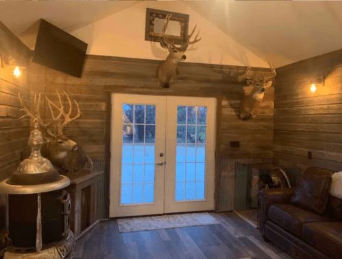 Cozy interior with wooden walls, deer mounts, a TV, and a leather couch near double doors leading outside.