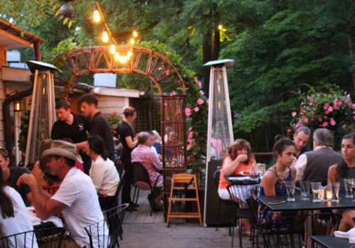 A lively outdoor restaurant scene with diners at tables, string lights, and greenery in the background.