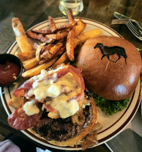 A gourmet burger topped with bacon and sauce, served with sweet potato fries and a bun featuring a horse logo.