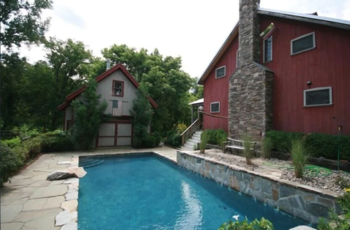 A red barn-style house with a stone chimney beside a blue swimming pool and lush greenery.