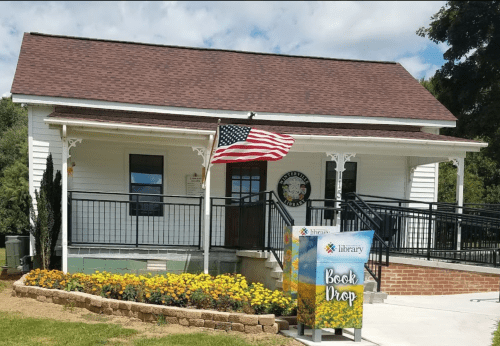 A small library building with an American flag, a sign for book drop, and colorful flowers in front.
