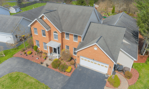 Aerial view of a large brick house with a landscaped yard and driveway, surrounded by trees and other homes.