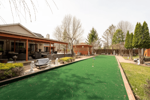 A backyard with a green lawn, patio area, and trees, featuring a small putting green and outdoor seating.