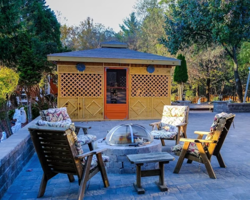 A cozy outdoor seating area with two chairs and a fire pit in front of a wooden gazebo surrounded by trees.