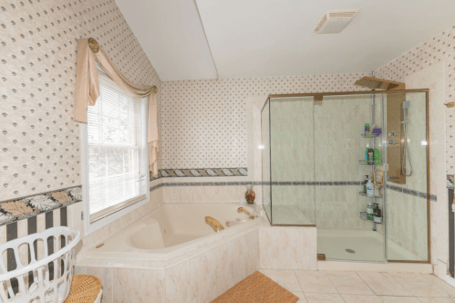 A spacious bathroom featuring a corner bathtub, glass shower, and patterned wallpaper. Natural light from a window.