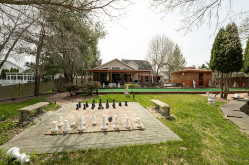 A backyard with a large outdoor chess set, benches, and a house in the background surrounded by trees.
